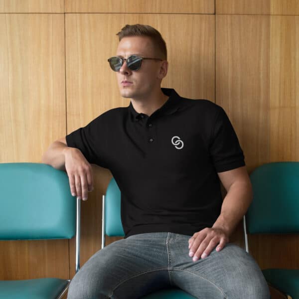 A person wearing men's sunglasses, a Men's Premium Polo with a small logo on the chest, and jeans is sitting on a blue chair against a wooden wall. They have a serious expression and are resting one arm on the chair beside them.