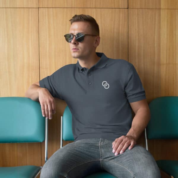 A man wearing premium sunglasses and a gray Men's Premium Polo with a circular logo on it sits casually on a green chair against a wooden wall. He rests one arm on the backrest of an adjacent chair, looking off to the side.